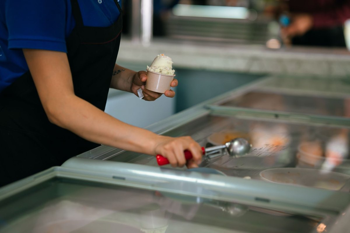 A worker in an apron scooping ice cream into a paper cup at an open freezer.